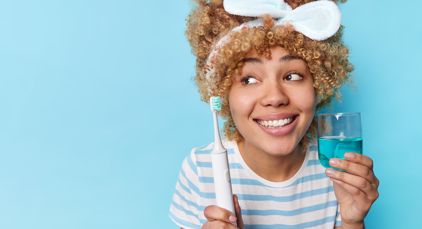 Person with curly hair wearing a headband, holding a toothbrush and glass of blue liquid against a light blue background